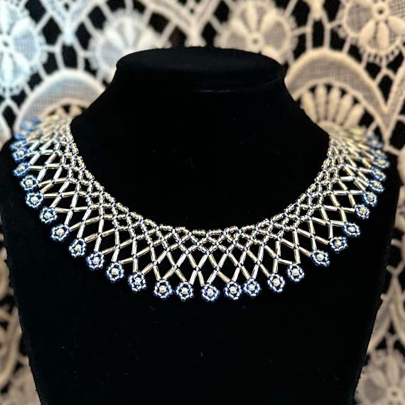 A photo of a blue and silver lace collar necklace on a velvet stand with a white lace background.